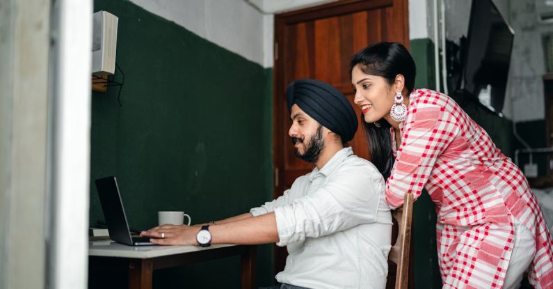 Desk - Side view of positive young Sikh man in shirt and turban working on laptop at home while wife leaning on chair behind